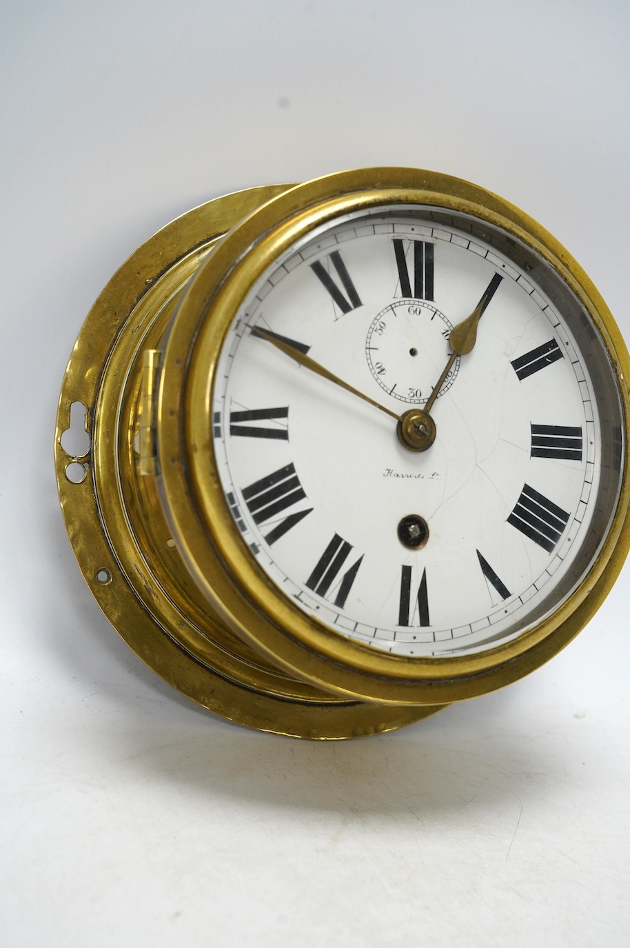 An early 20th century brass bulkhead timepiece retailed by Harrods, dial 13.5cm. Condition - poor to fair, seconds hand missing and some cracks to enamel dial